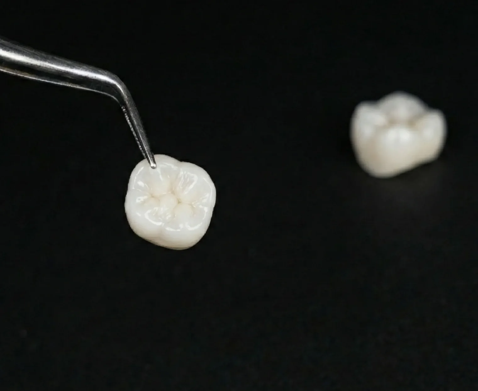 A dental crown held in tweezers, a second crown beside it, on black