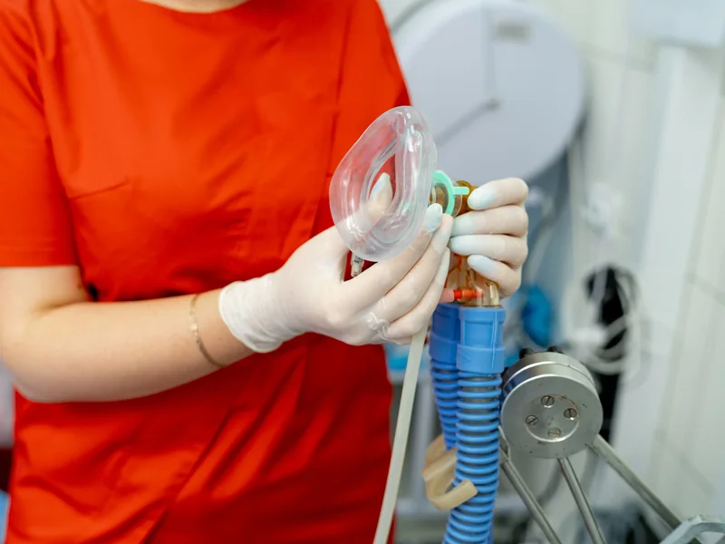 A clinician in gloves holding an anesthesia mask and breathing circuit